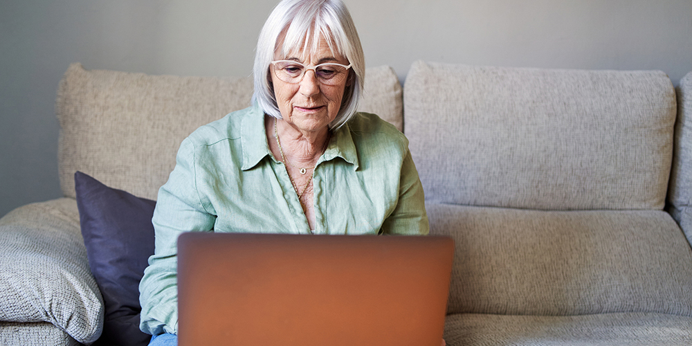 An elderly woman with white hair and glasses sits on a beige couch, focused on using a laptop placed in front of her, in a calm indoor setting